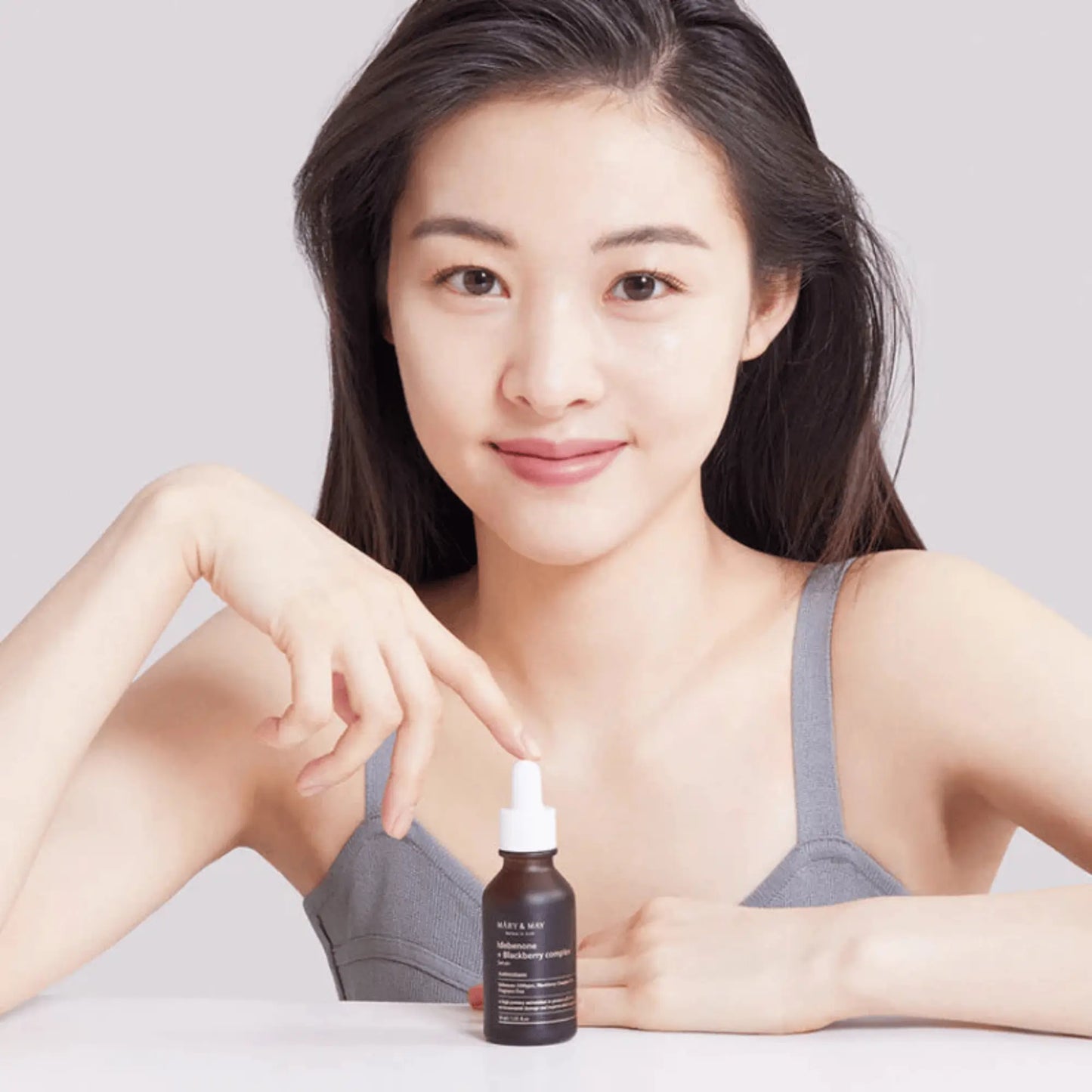 Mary & May Idebenone + Blackberry Complex Serum 30ml, sérum facial coreano antioxidante que previne envelhecimento precoce, melhora firmeza e dá vitalidade, modelo feminina a segurar o frasco junto ao rosto sobre fundo branco, cosmética coreana skincare k-beauty Portugal.