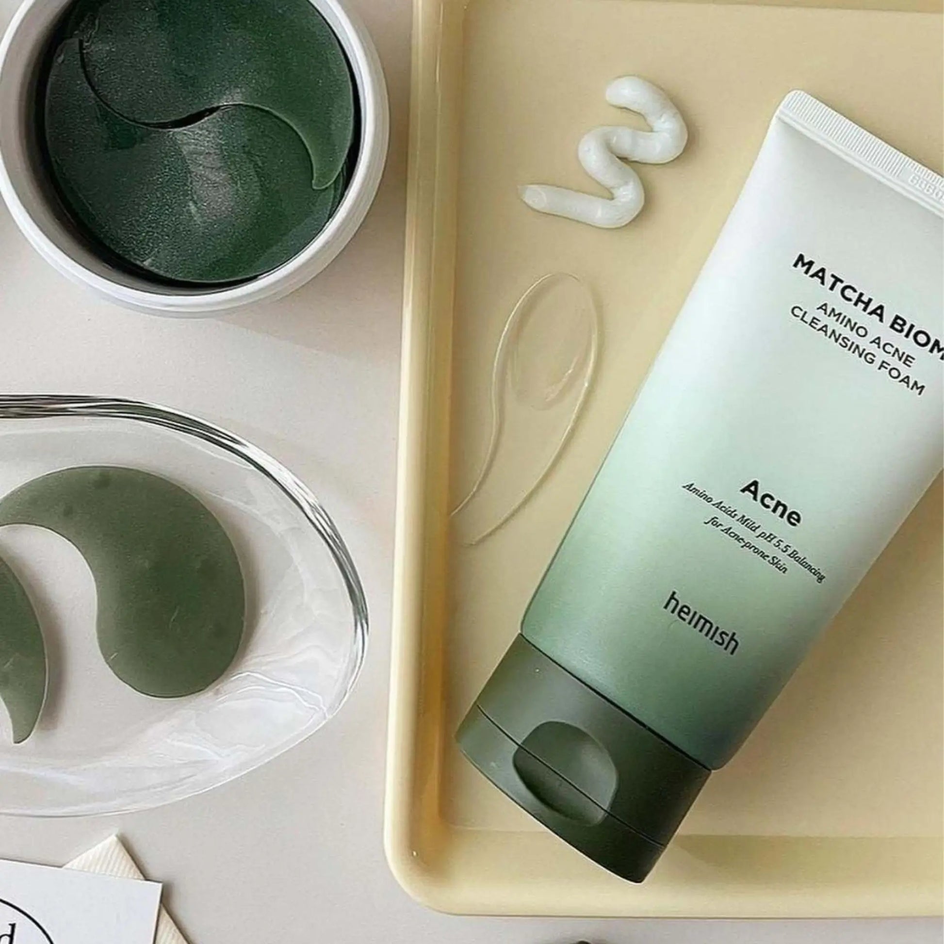 Heimish Matcha Biome Amino Acne Cleansing Foam 150g, espuma de limpeza facial coreana que remove impurezas, controla oleosidade e acalma irritações, frasco em pé com acessórios de skincare e pó de matcha ao lado sobre fundo bege, cosmética coreana skincare k-beauty Portugal.