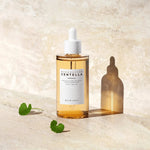 Madagascar Centella Ampoule da Skin1004 em frasco grande com folhas de centella ao lado sobre mármore bege