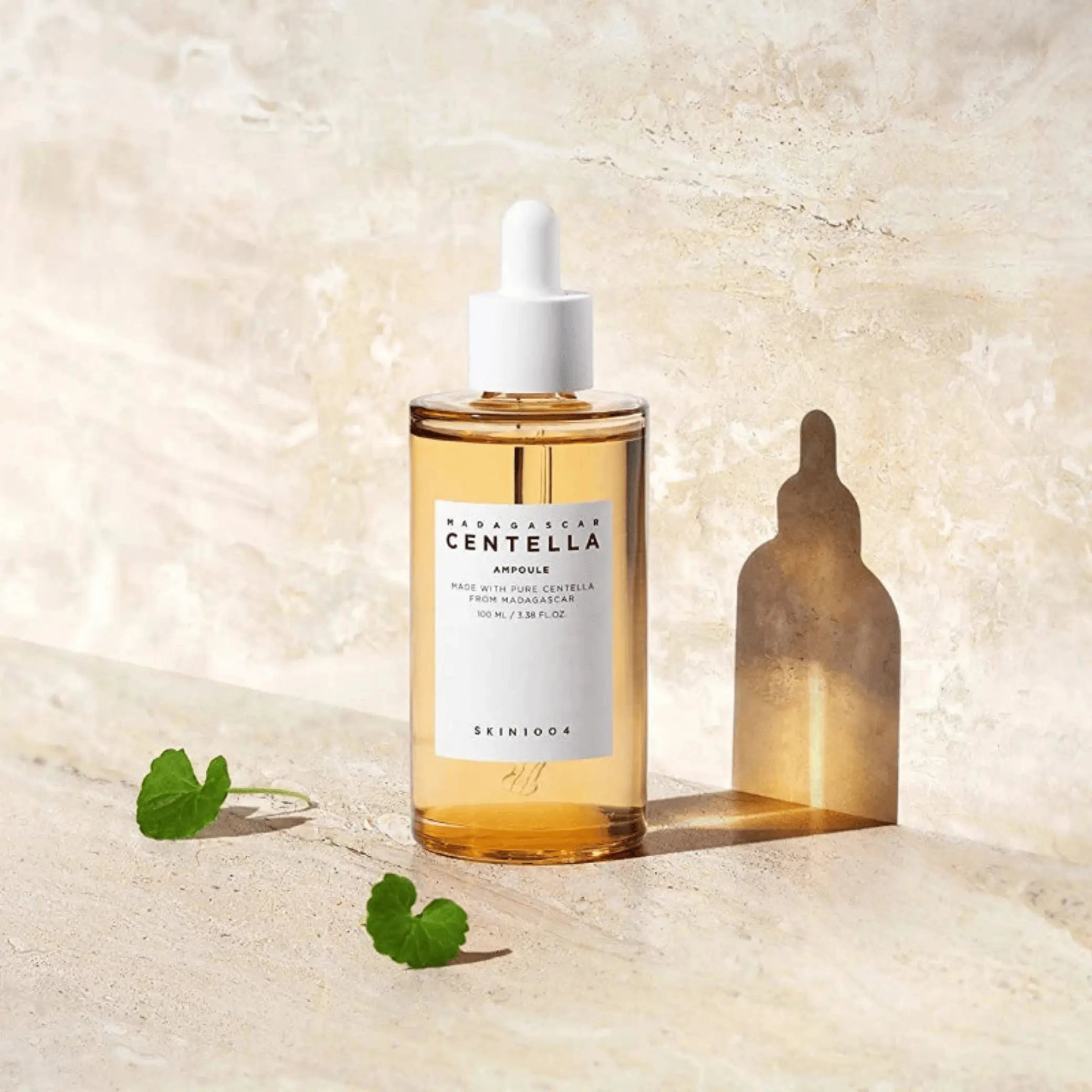 Madagascar Centella Ampoule da Skin1004 em frasco grande com folhas de centella ao lado sobre mármore bege